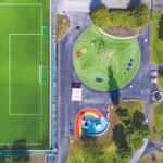 Aerial view of a sports field on the left and a park playground on the right, with a climbing net, rainbow slide, and green, circular play area.