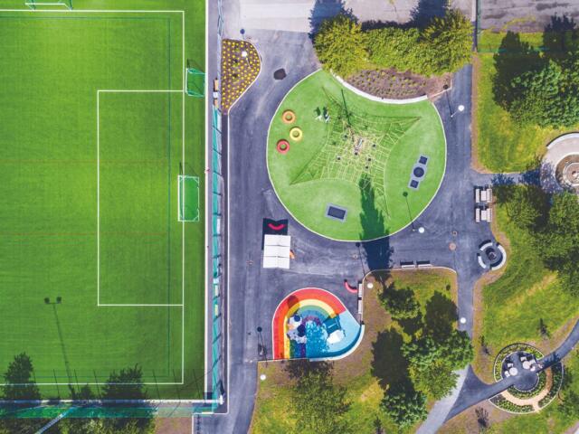 Aerial view of a sports field on the left and a park playground on the right, with a climbing net, rainbow slide, and green, circular play area.