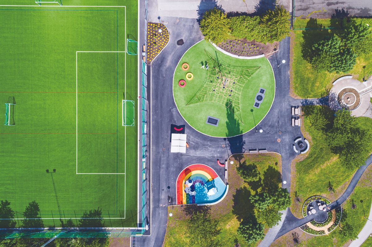 Aerial view of a sports field on the left and a park playground on the right, with a climbing net, rainbow slide, and green, circular play area.