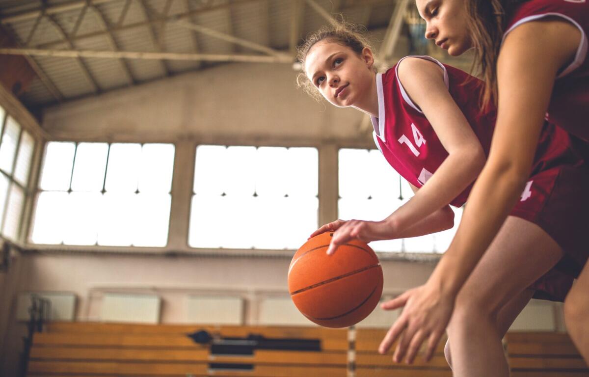 Two girls in red basketball jerseys bend over and dribble an orange basketball on an indoor court, focused on the play for a game or drill in progress inside a gym.