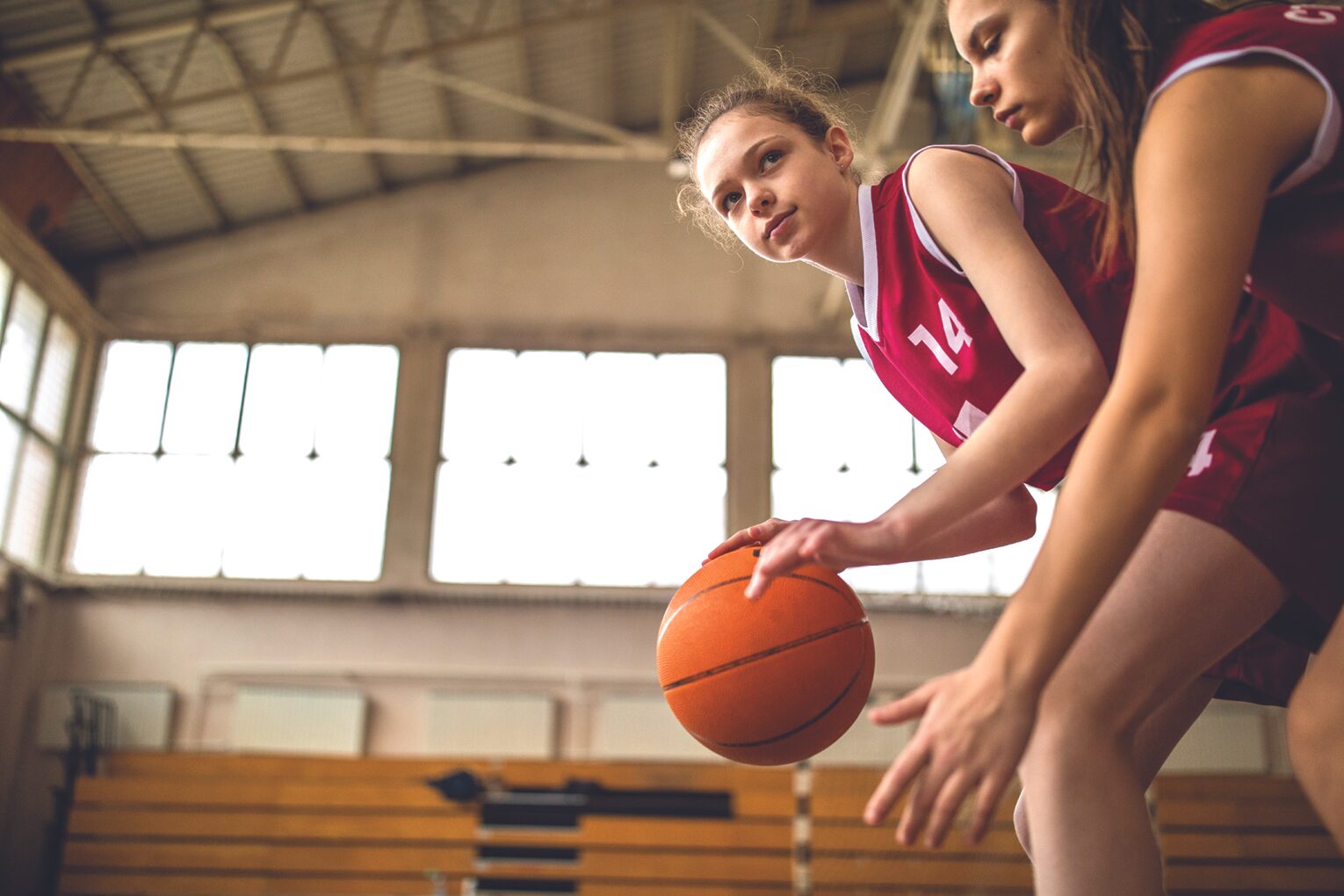 Two girls in red basketball jerseys bend over and dribble an orange basketball on an indoor court, focused on the play for a game or drill in progress inside a gym.