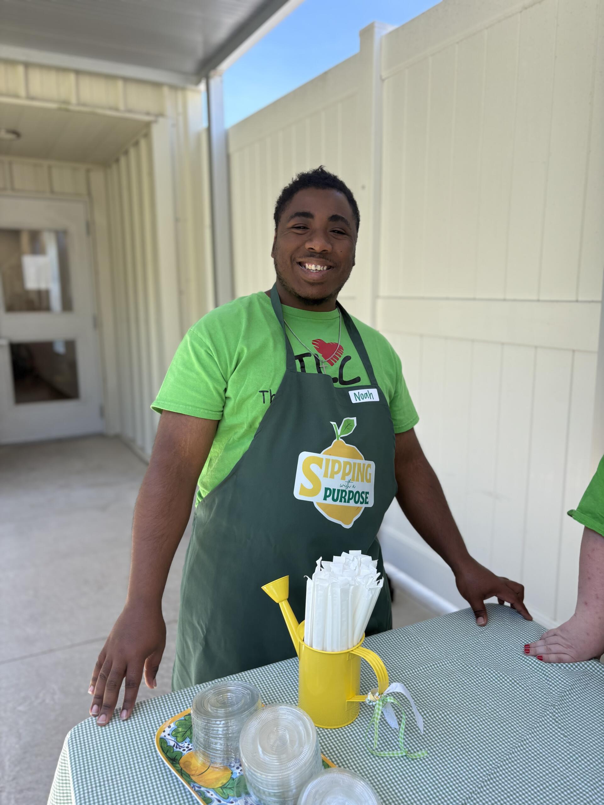 Smiling man in a green shirt and apron stands behind a table with a yellow pitcher and stacked cups outdoors.