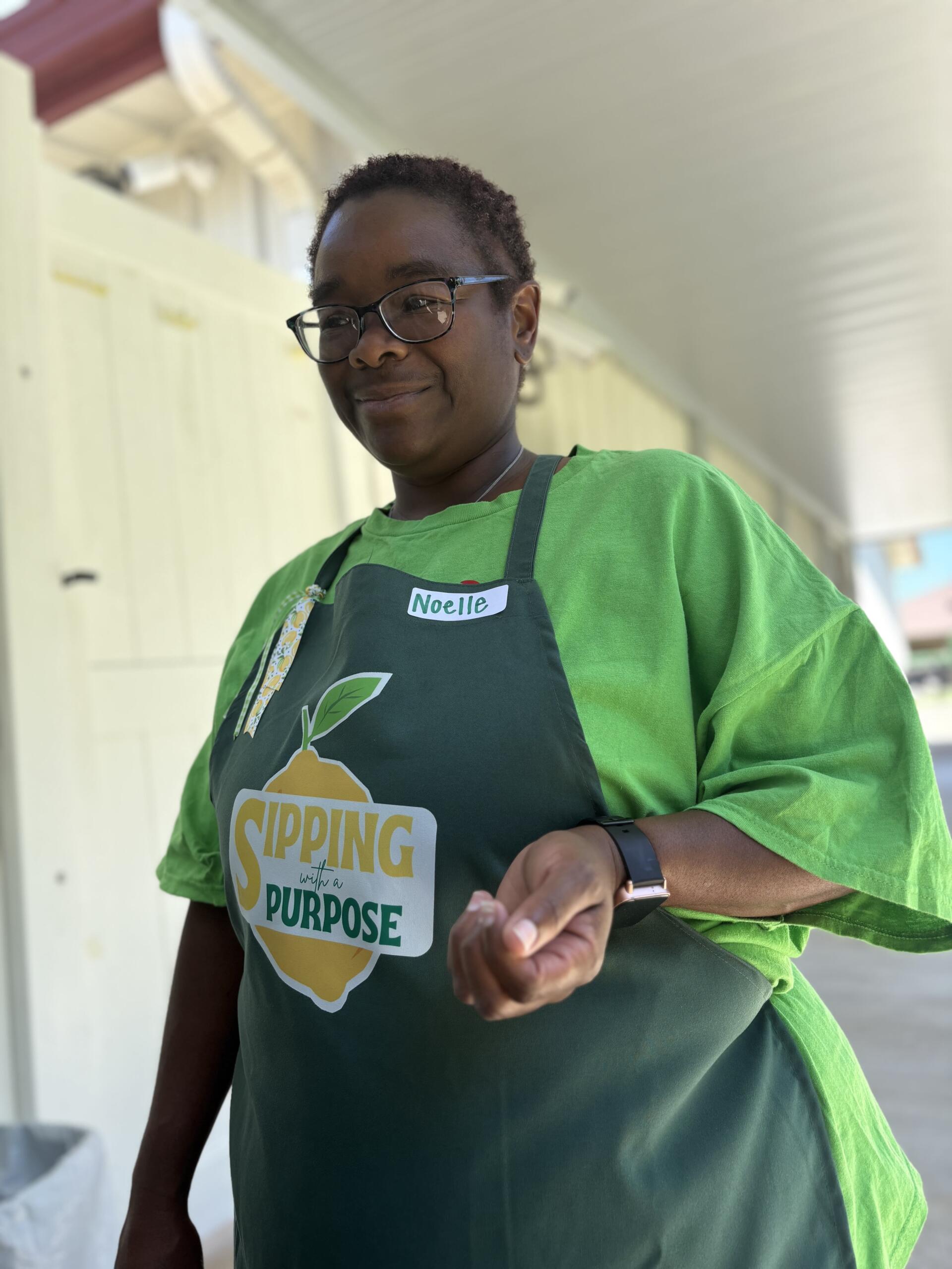 Noelle in a green shirt and dark apron with a 'Sipping with a Purpose' logo, standing outdoors and smiling.