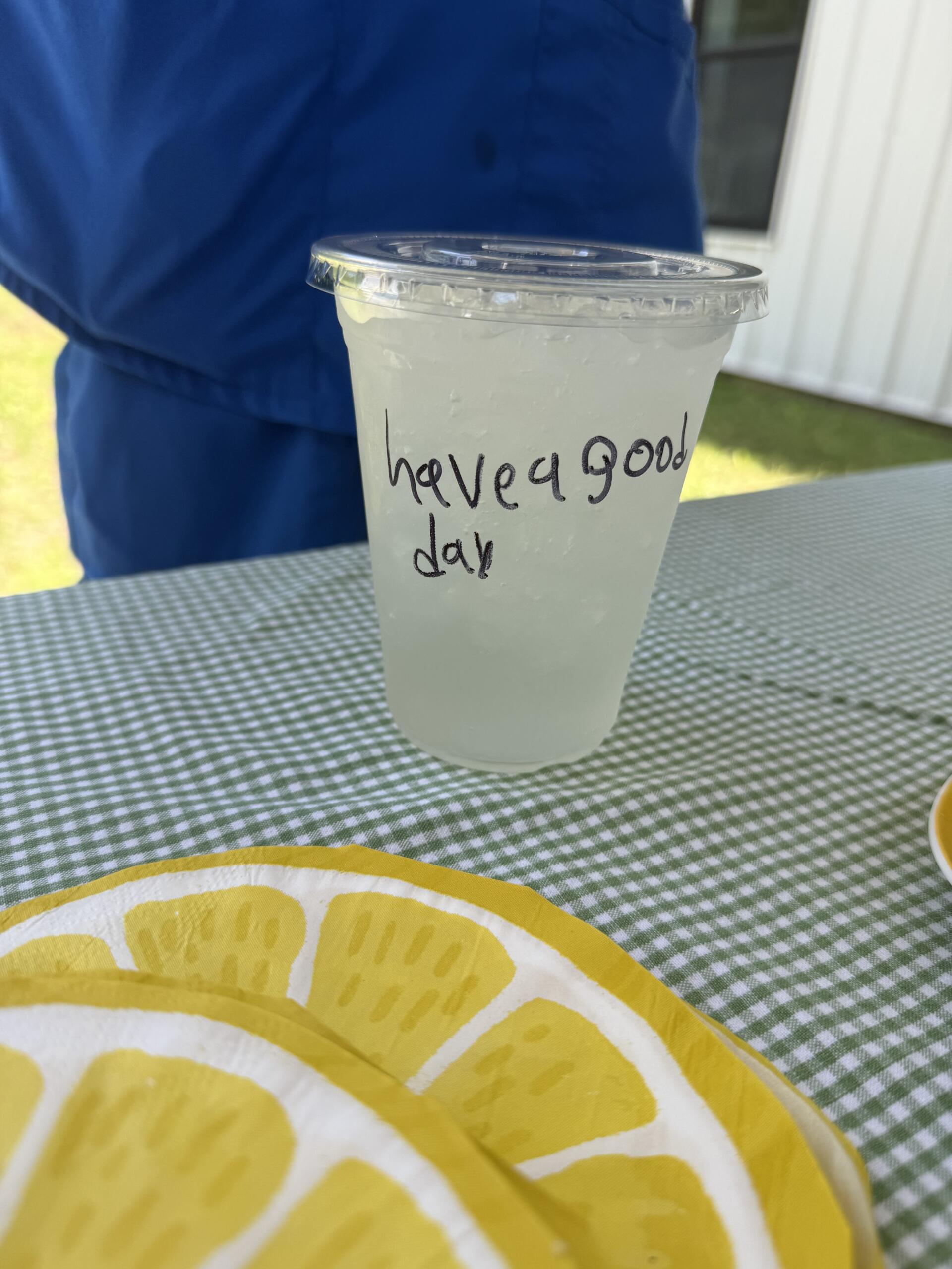 Plastic cup with ice and a lid, resting on a checkered outdoor table; handwritten message on the cup reads 'have a good day.'