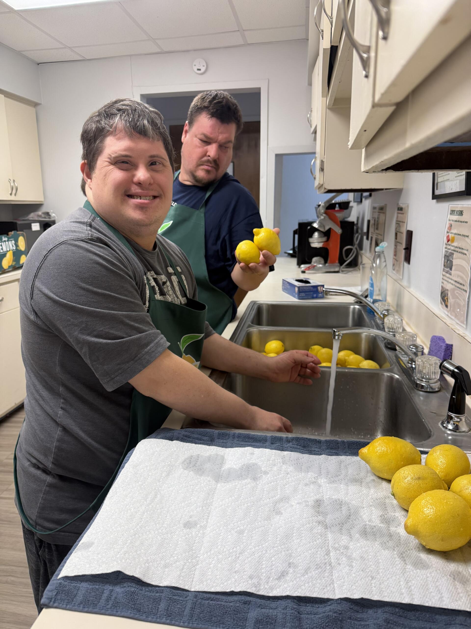 Two men in a kitchen wash lemons at a double sink; one smiles at the camera while the other holds a lemon and looks on.