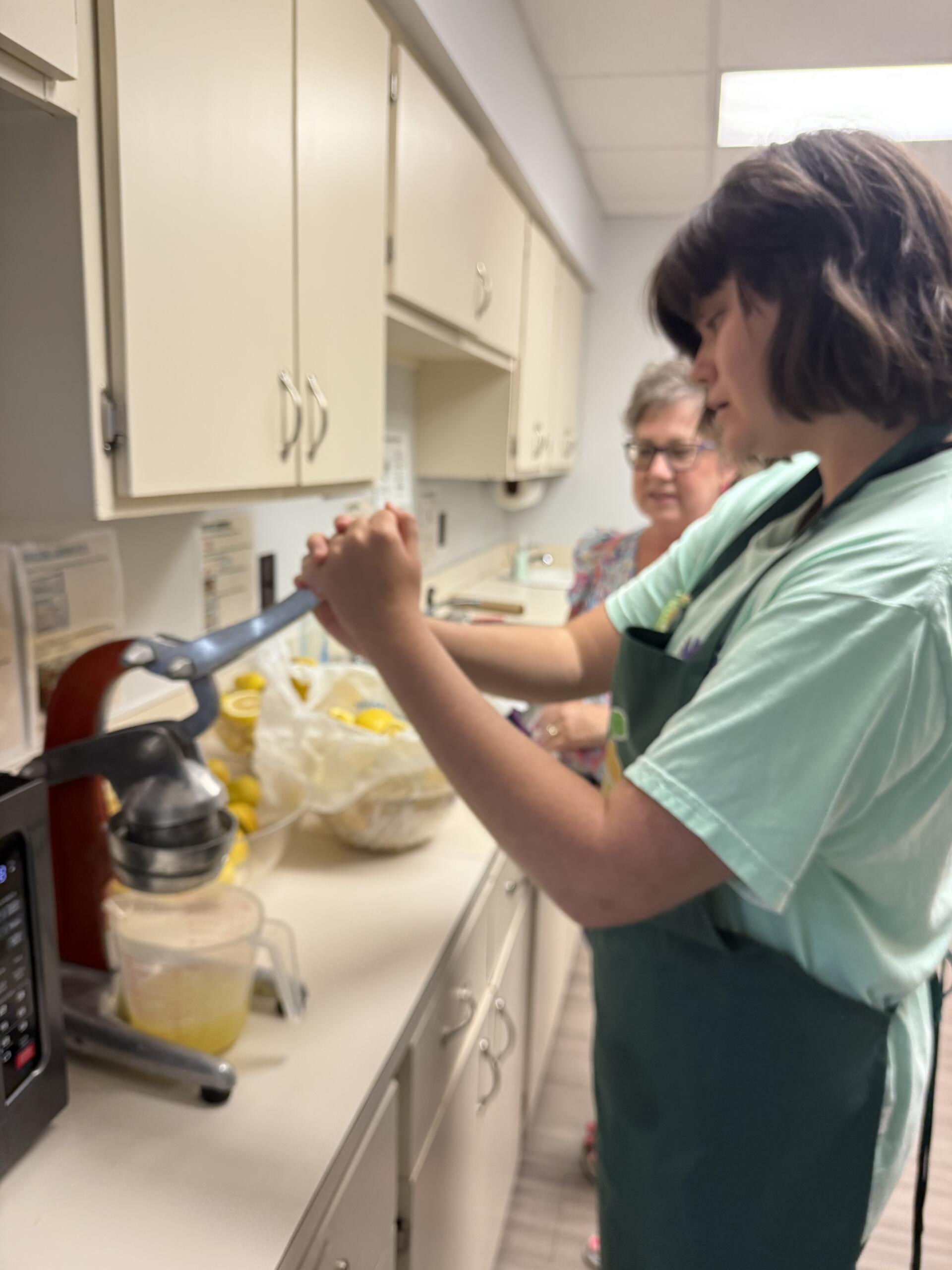 Healthcare worker in teal scrubs at a kitchen counter, measuring or pouring liquid, with another person in the background.
