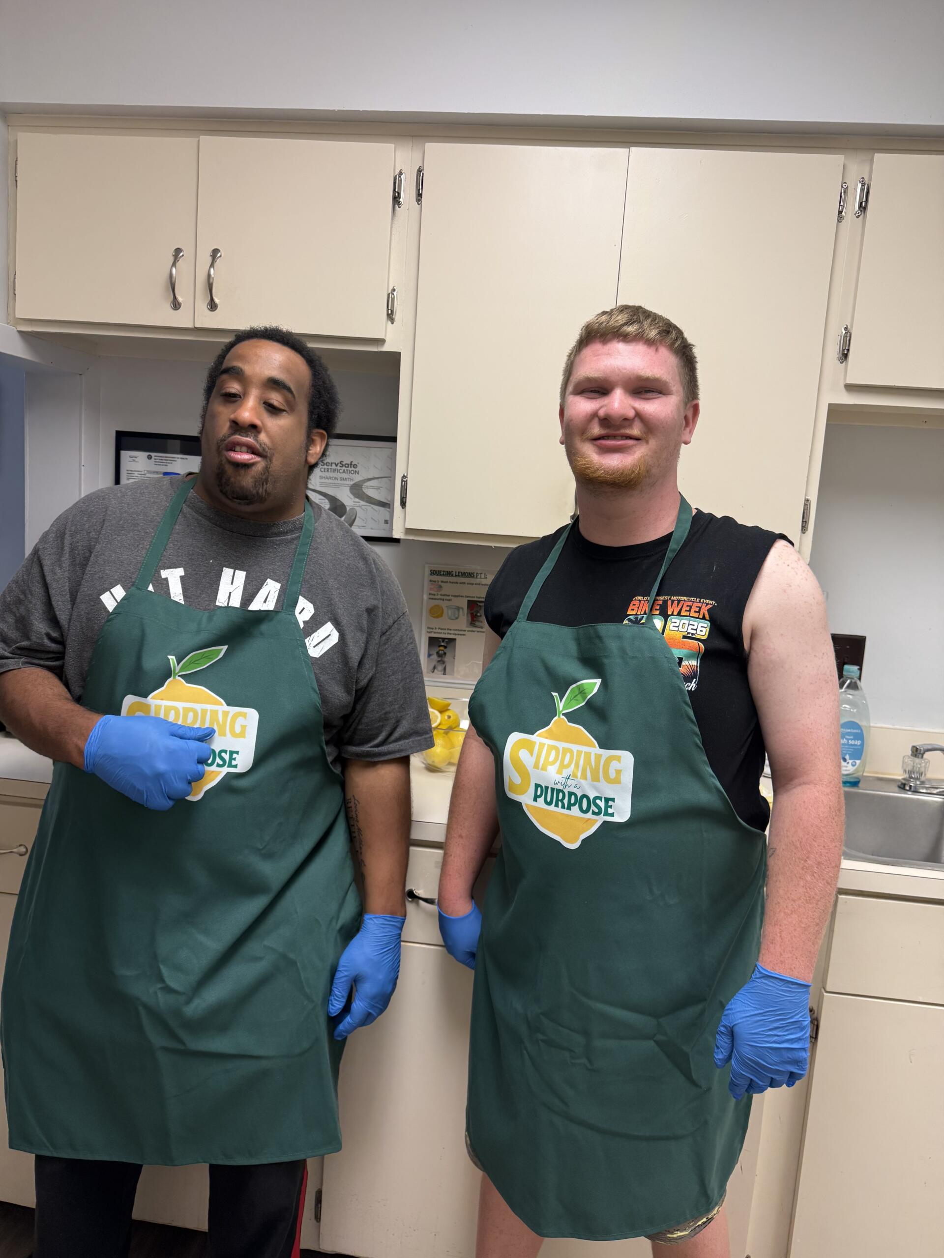 Two men in a kitchen wearing green aprons; one in a gray shirt with blue gloves, the other in a black sleeveless shirt, both smiling at the camera.