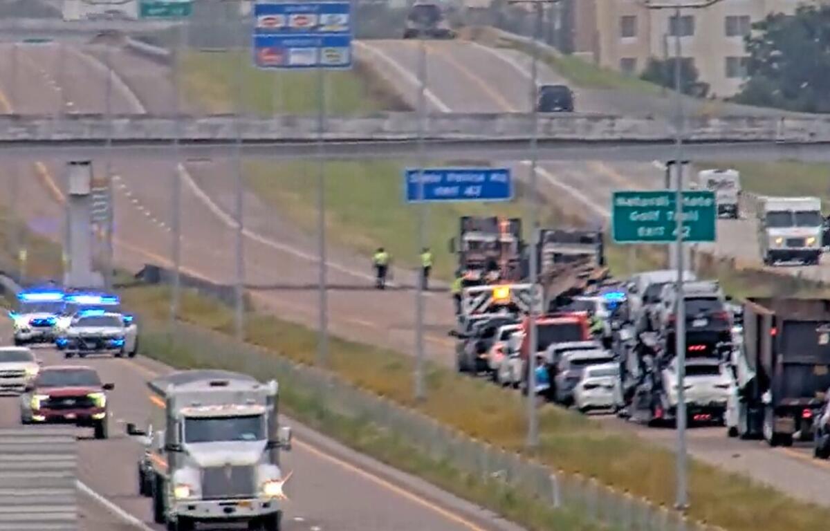 Highway emergency scene with blue flashing lights, multiple vehicles stopped along the right shoulder and responders near a truck on a congested roadway under overpass signs.