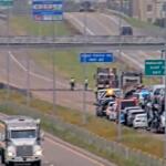 Highway emergency scene with blue flashing lights, multiple vehicles stopped along the right shoulder and responders near a truck on a congested roadway under overpass signs.