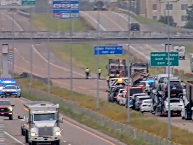 Highway emergency scene with blue flashing lights, multiple vehicles stopped along the right shoulder and responders near a truck on a congested roadway under overpass signs.