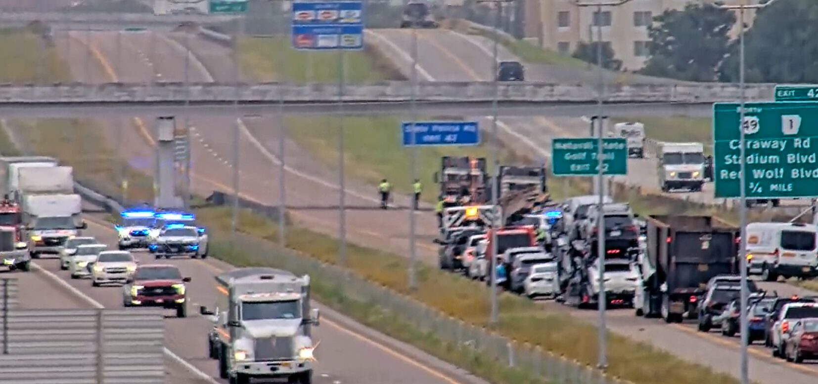 Highway emergency scene with blue flashing lights, multiple vehicles stopped along the right shoulder and responders near a truck on a congested roadway under overpass signs.