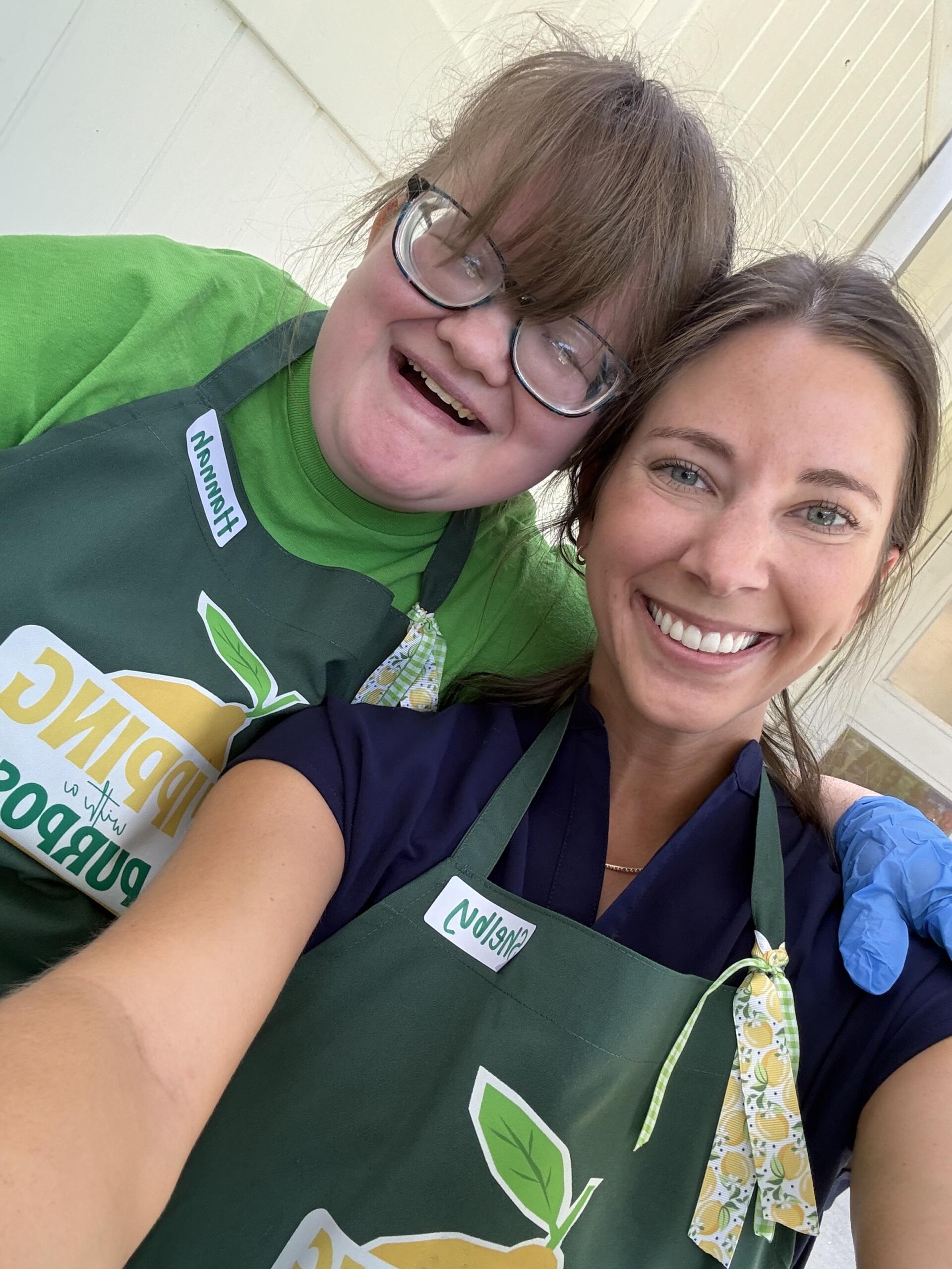 Two smiling people wearing green aprons pose for a selfie outdoors, with one person wearing glasses and bangs.