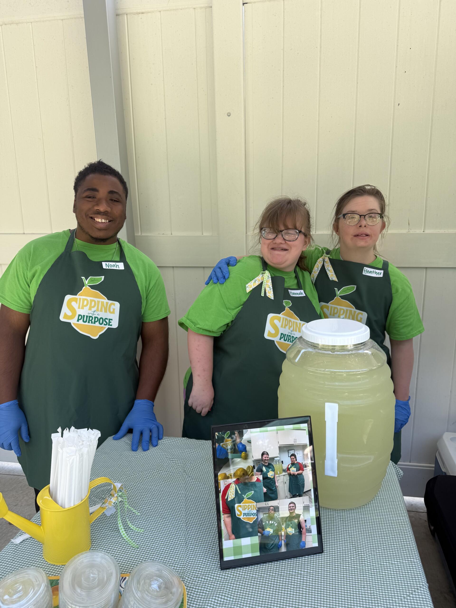 Three volunteers in green shirts and aprons pose at a lemonade stand, with a large jar of lemonade on the table and name tags visible (Noah, Hannah, Heather).