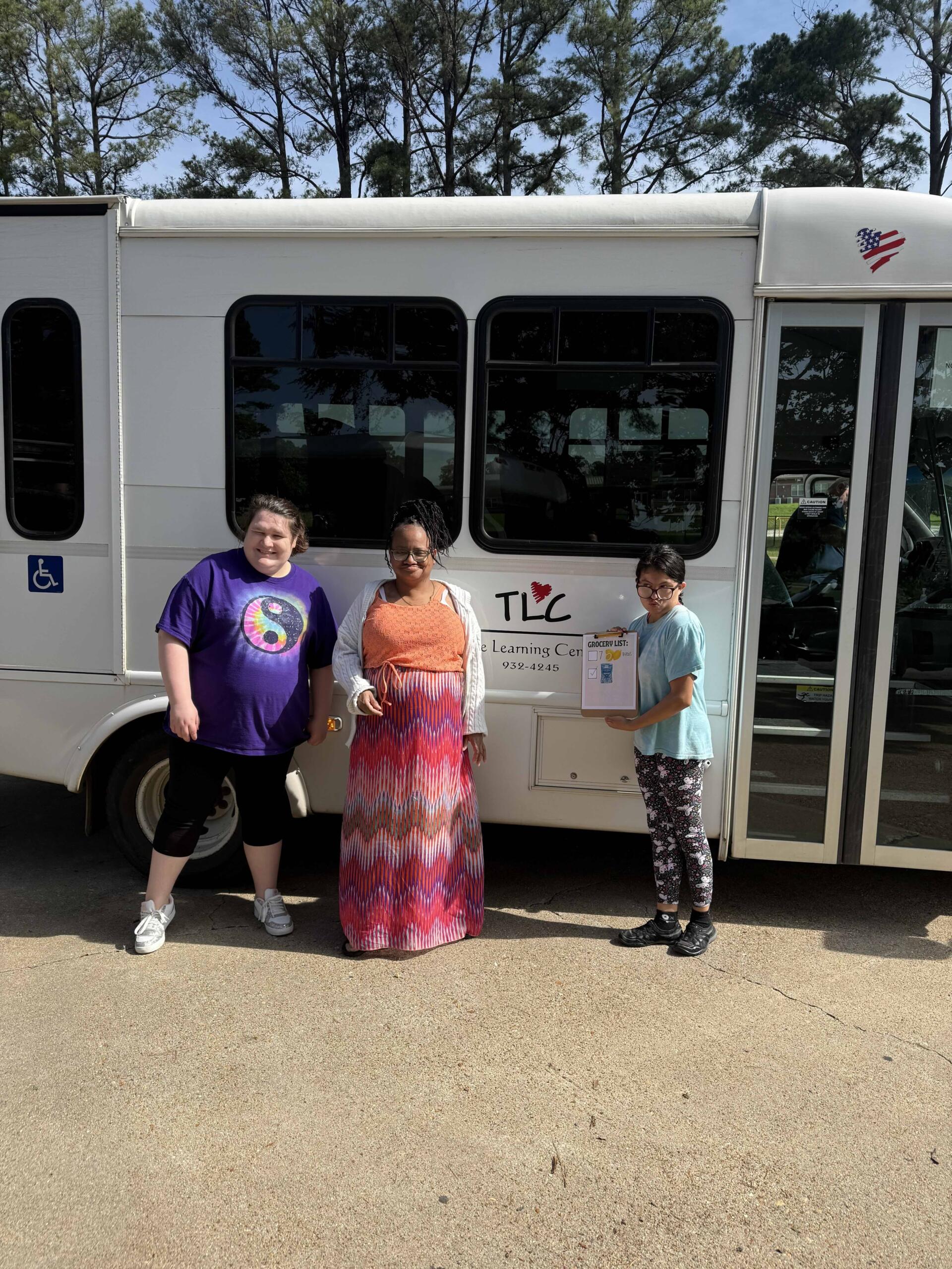 Three people stand in front of a white shuttle bus, with a TLC sign on the door and a clipboard held by the person on the right; trees are in the background.