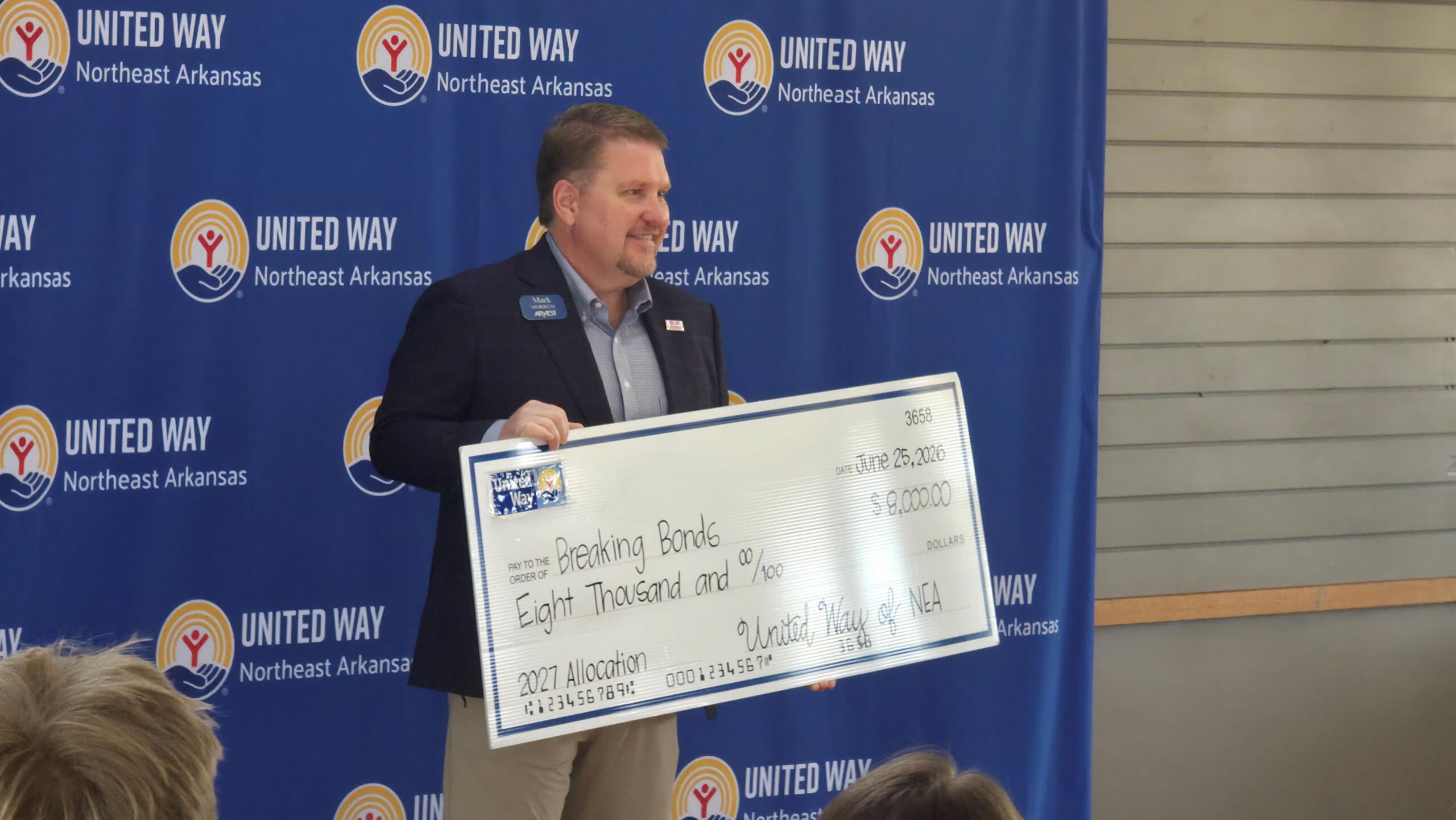 Man in a blazer holds a large ceremonial check at a United Way event, with a blue backdrop featuring United Way logos. (the check shows an amount of eight thousand dollars)