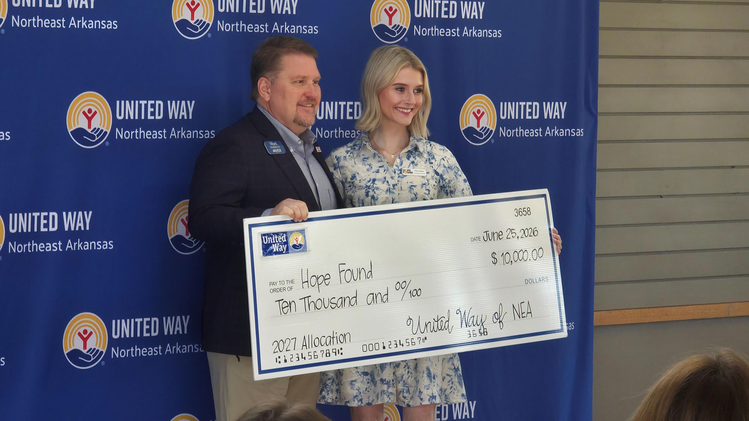 Two adults smiling while holding a large donation check in front of a United Way Northeast Arkansas backdrop showing the United Way logo.