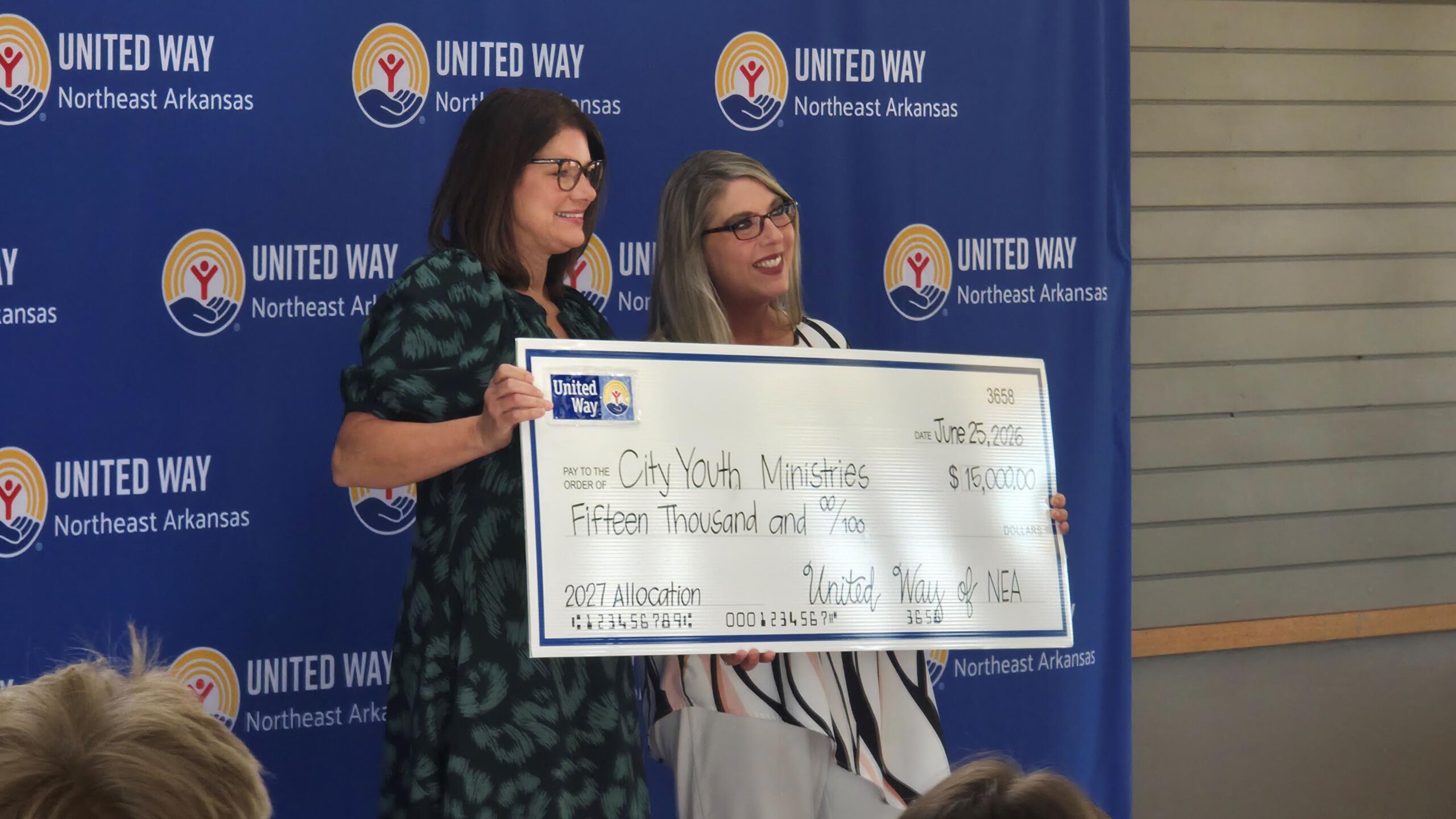 Two women stand in front of a United Way backdrop, holding a large ceremonial check for a donation act.