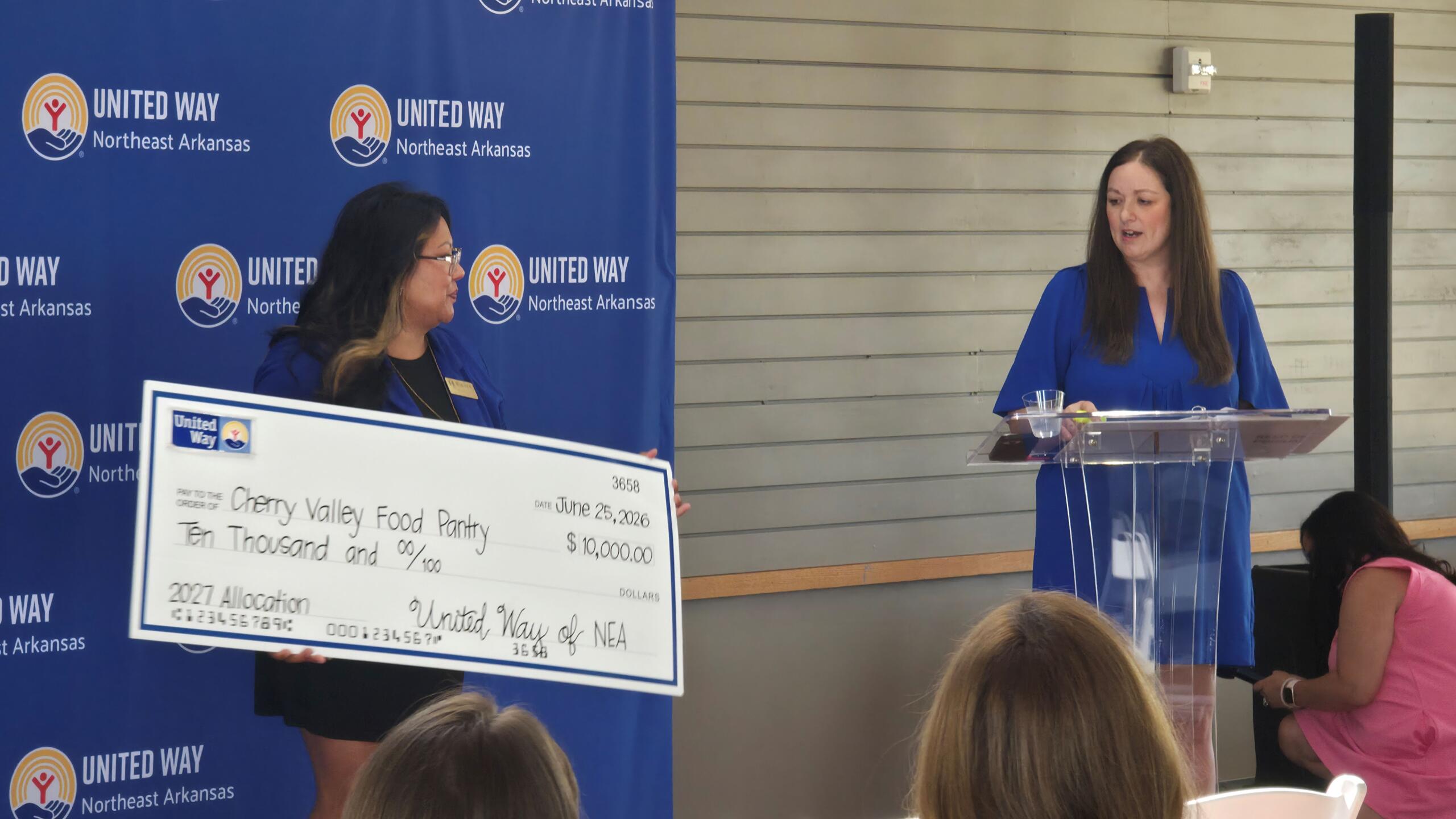 Two women at a United Way ceremony stand beside a large ceremonial check; one woman at left holds the check, others speak at a podium on the right.