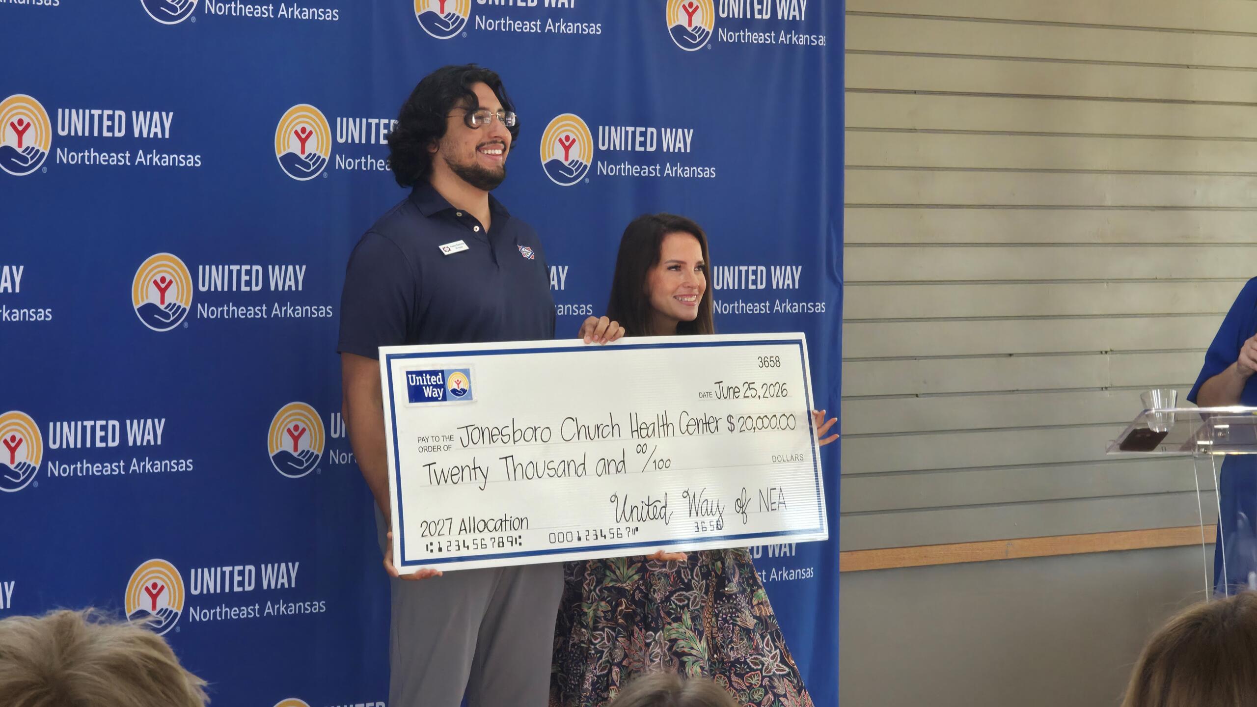 Two people stand on stage holding a large check from United Way NEA to Jonesboro Church Health Center for $20,000, with a blue United Way backdrop.