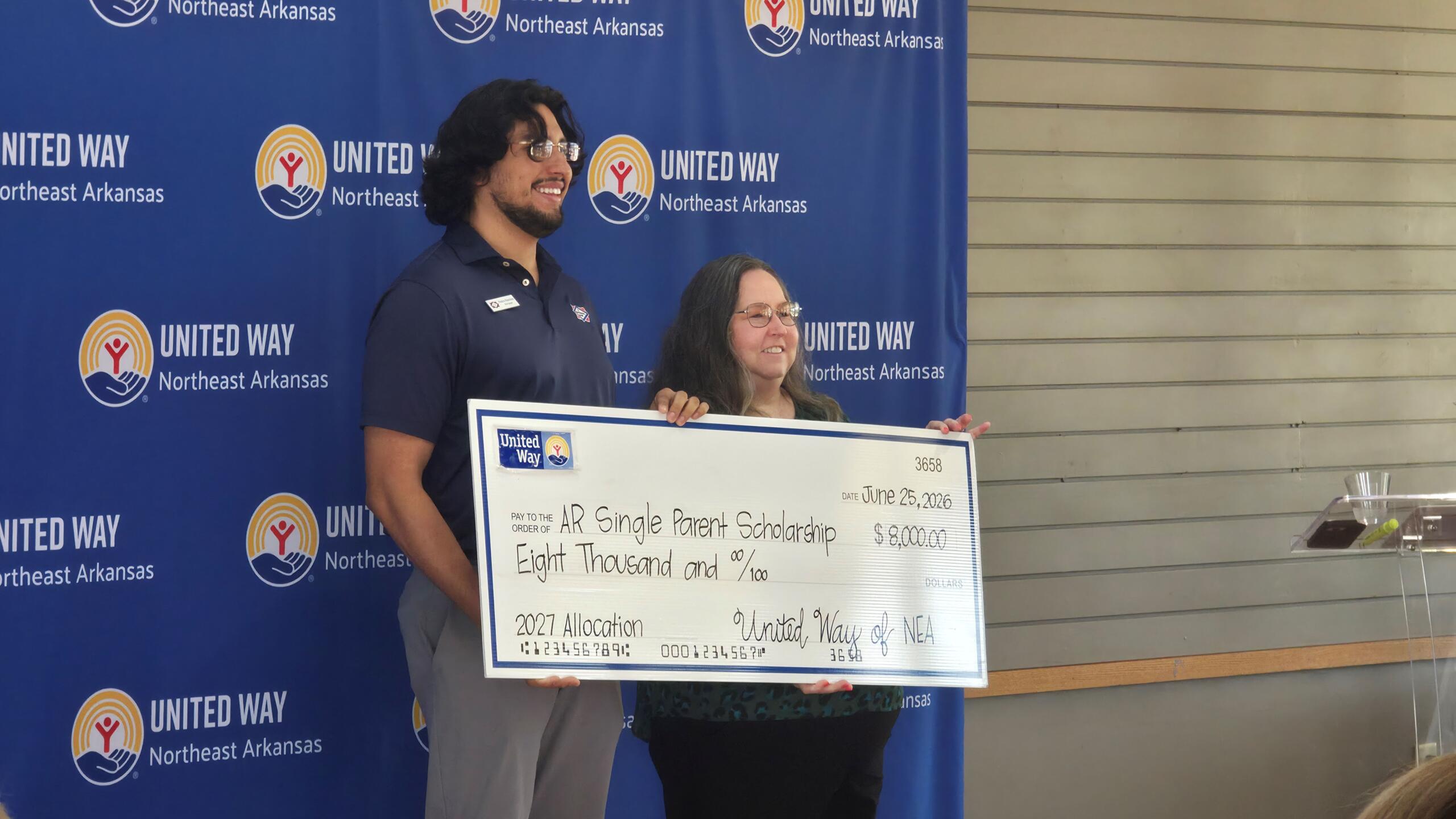 Two people stand side by side presenting a large check at a United Way event against a blue backdrop with Northeast Arkansas logos.