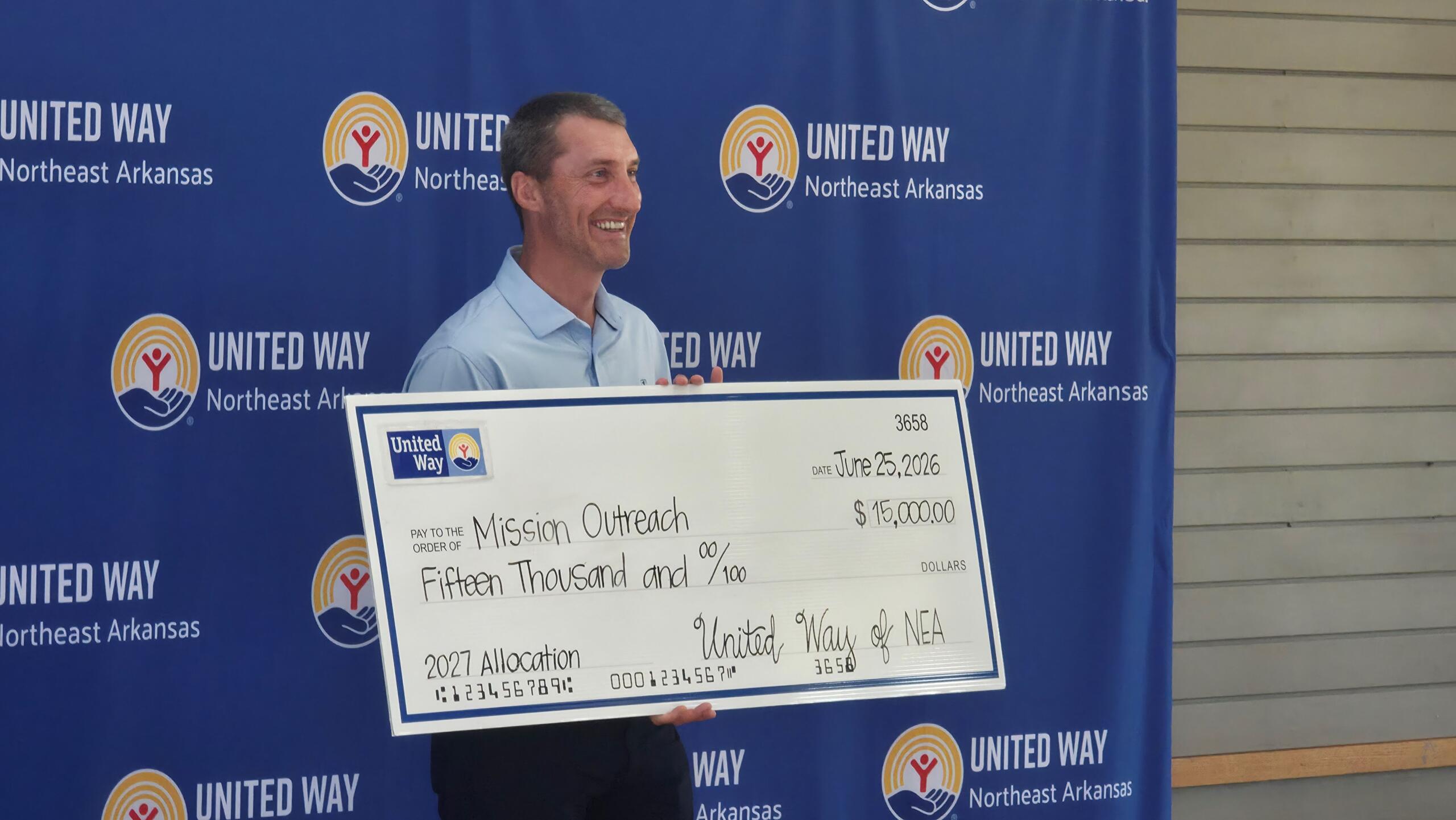 Smiling man in a light blue shirt holds a large check at a United Way event with a United Way backdrop behind him (Northeast Arkansas).