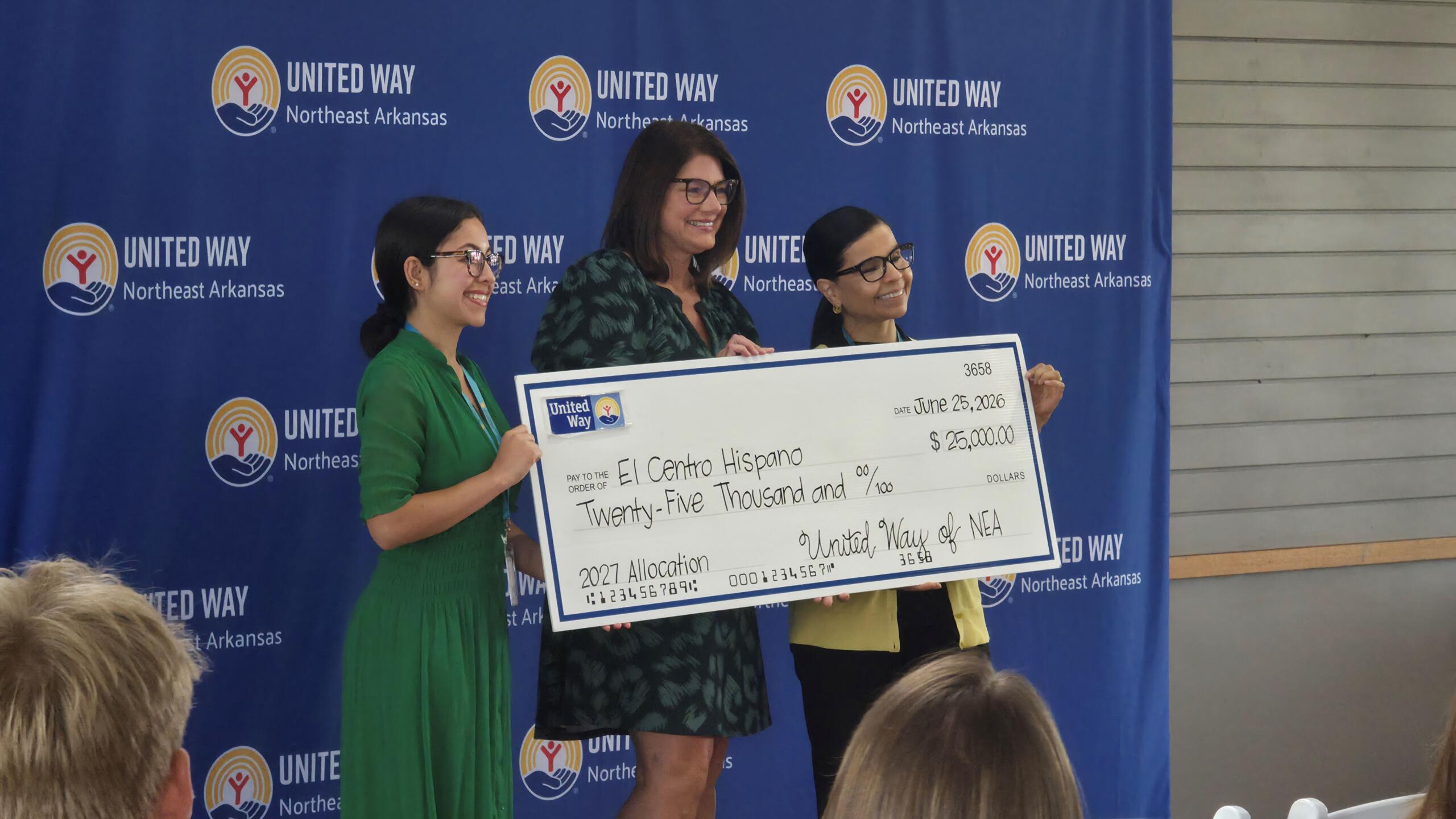 Three women hold a large ceremonial check at a United Way event in front of a blue United Way backdrop with logos.