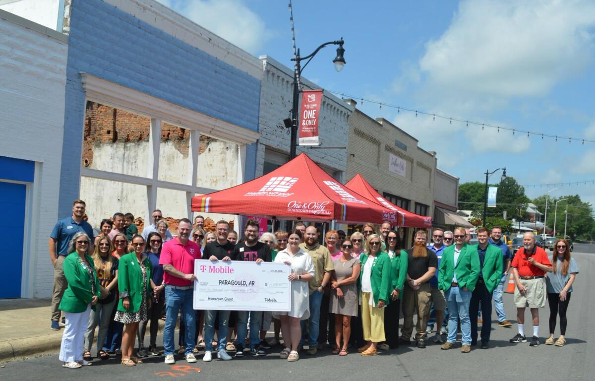 Large group of people posing on a street with a big ceremonial check for a hometown grant in Paragould, Arkansas, under a red canopy.