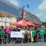 Large group of people posing on a street with a big ceremonial check for a hometown grant in Paragould, Arkansas, under a red canopy.