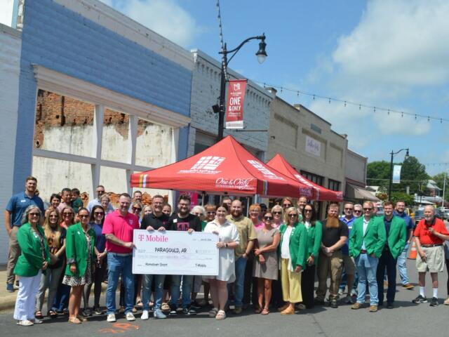 Large group of people posing on a street with a big ceremonial check for a hometown grant in Paragould, Arkansas, under a red canopy.