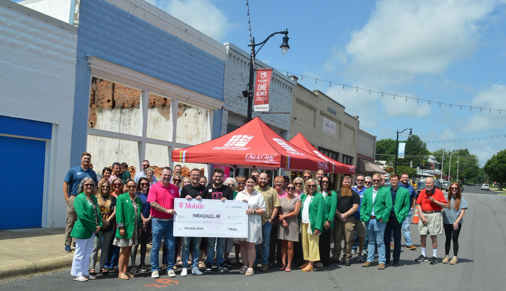 Large group of people posing on a street with a big ceremonial check for a hometown grant in Paragould, Arkansas, under a red canopy.