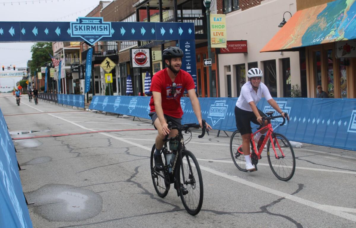 Cyclists ride through a closed city street with blue race barriers and a large overhead banner reading 'Skirmish' festival.