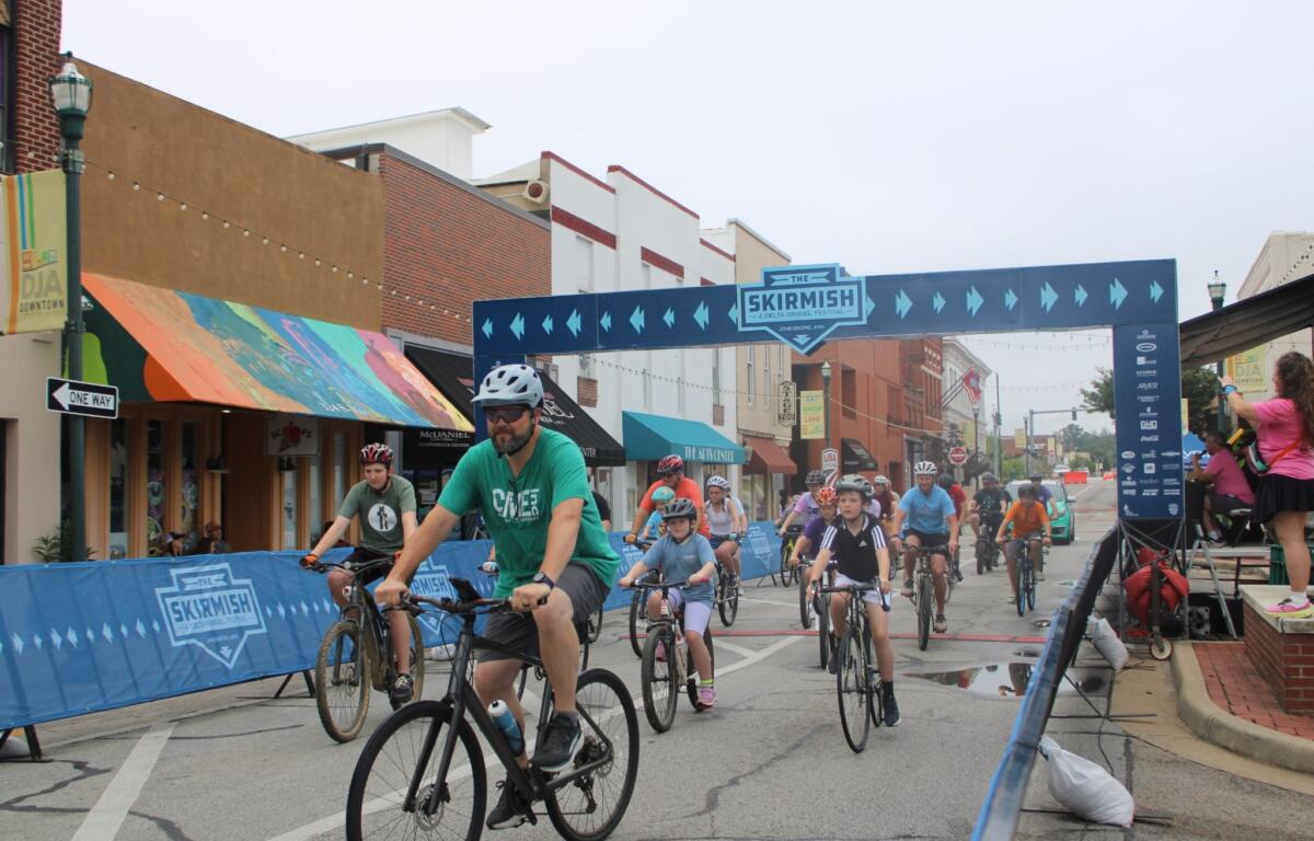 Bicyclists race down a city street under a blue finish banner that says The Skirmish.