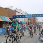 Bicyclists race down a city street under a blue finish banner that says The Skirmish.