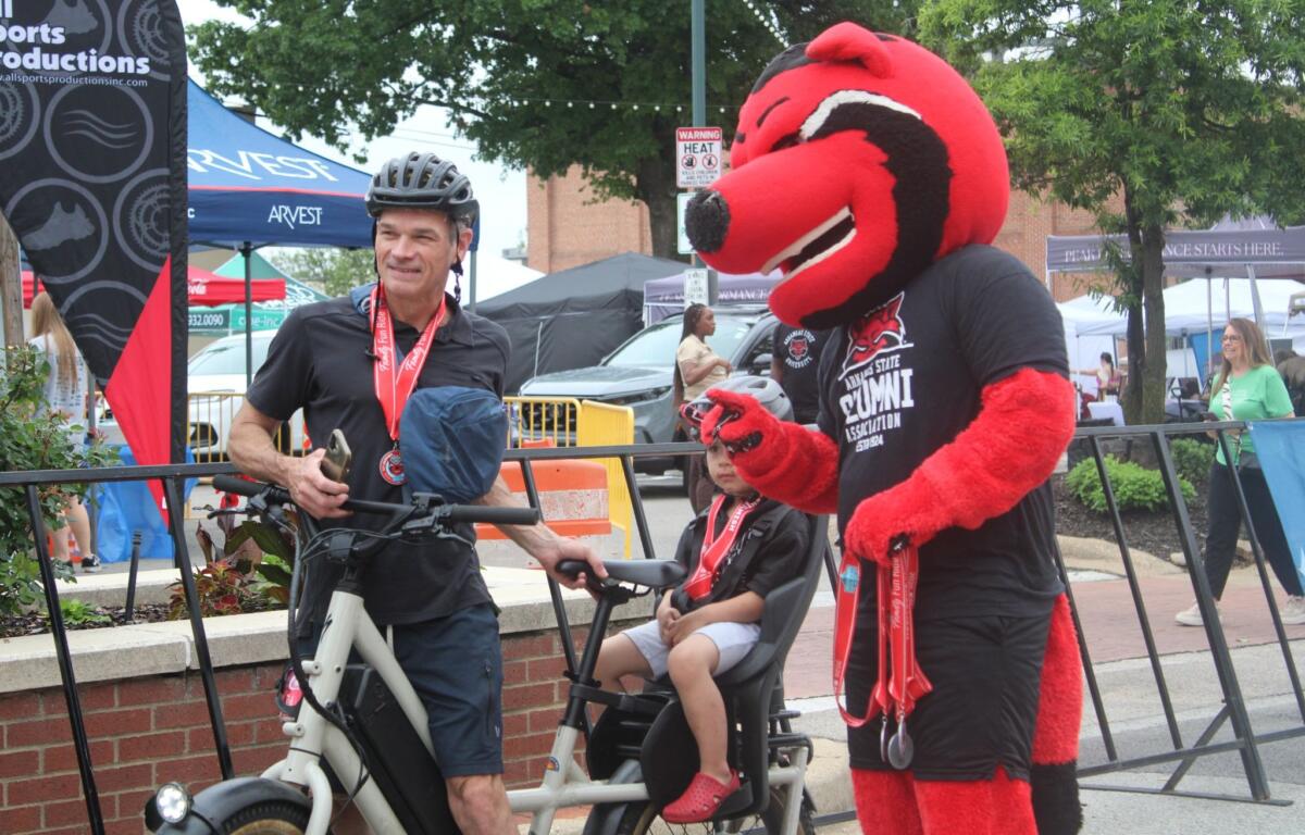 Man wearing a bike helmet standing next to a child in a stroller at an outdoor festival with tents in the background and a red mascot nearby.