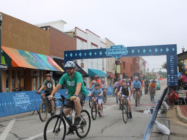 Bicyclists race down a city street under a blue finish banner that says The Skirmish.