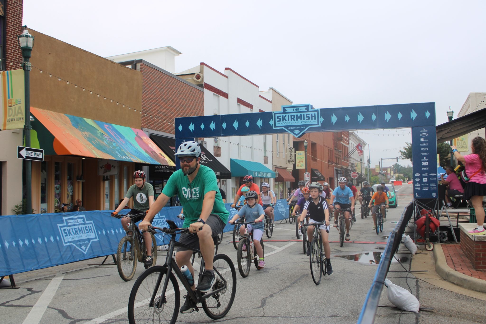 Bicyclists race down a city street under a blue finish banner that says The Skirmish.