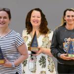 Three women stand side by side smiling, each holding a blue triangular trophy with a wooden base against a plain gray background. Left: striped shirt; middle: floral dress; right: dark sweatshirt with a tiger graphic.