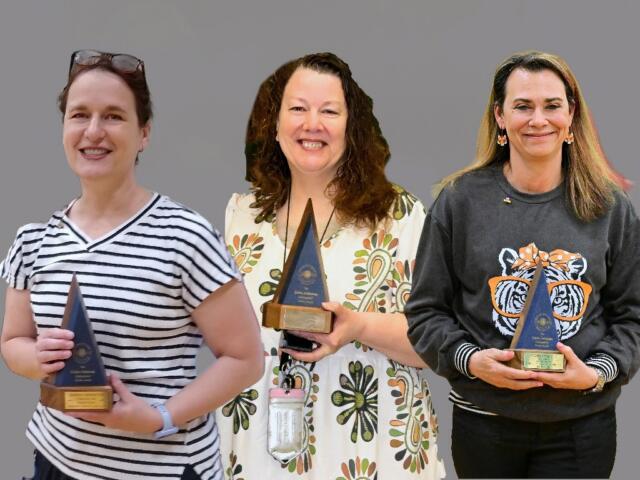 Three women stand side by side smiling, each holding a blue triangular trophy with a wooden base against a plain gray background. Left: striped shirt; middle: floral dress; right: dark sweatshirt with a tiger graphic.