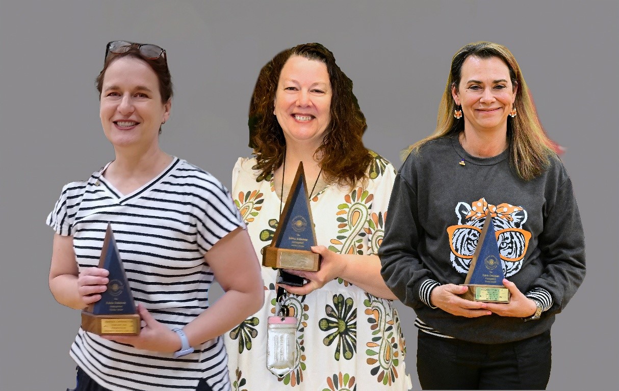 Three women stand side by side smiling, each holding a blue triangular trophy with a wooden base against a plain gray background. Left: striped shirt; middle: floral dress; right: dark sweatshirt with a tiger graphic.