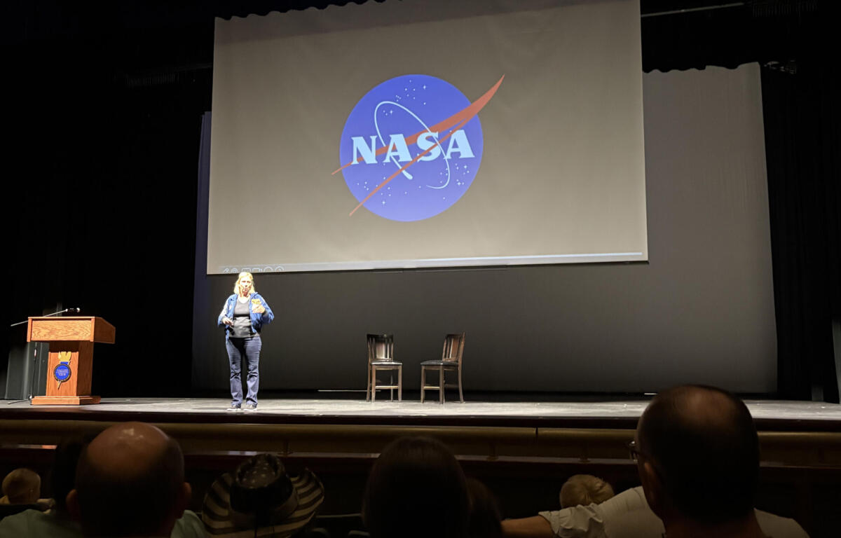 Presenter on stage in front of a large screen displaying the NASA logo; audience seated in an auditorium.