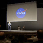 Presenter on stage in front of a large screen displaying the NASA logo; audience seated in an auditorium.