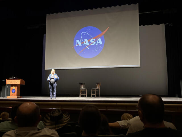 Presenter on stage in front of a large screen displaying the NASA logo; audience seated in an auditorium.
