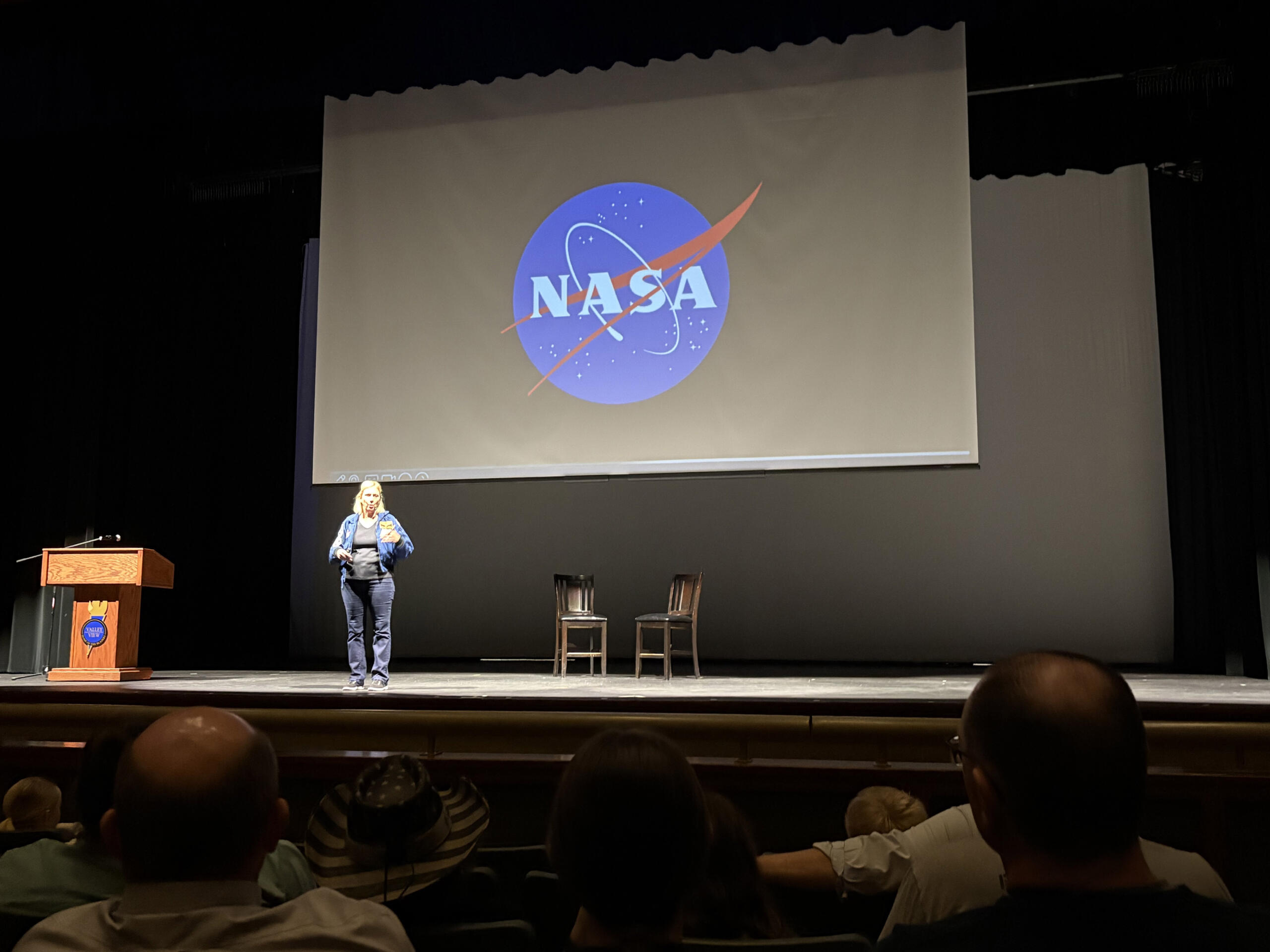 Presenter on stage in front of a large screen displaying the NASA logo; audience seated in an auditorium.