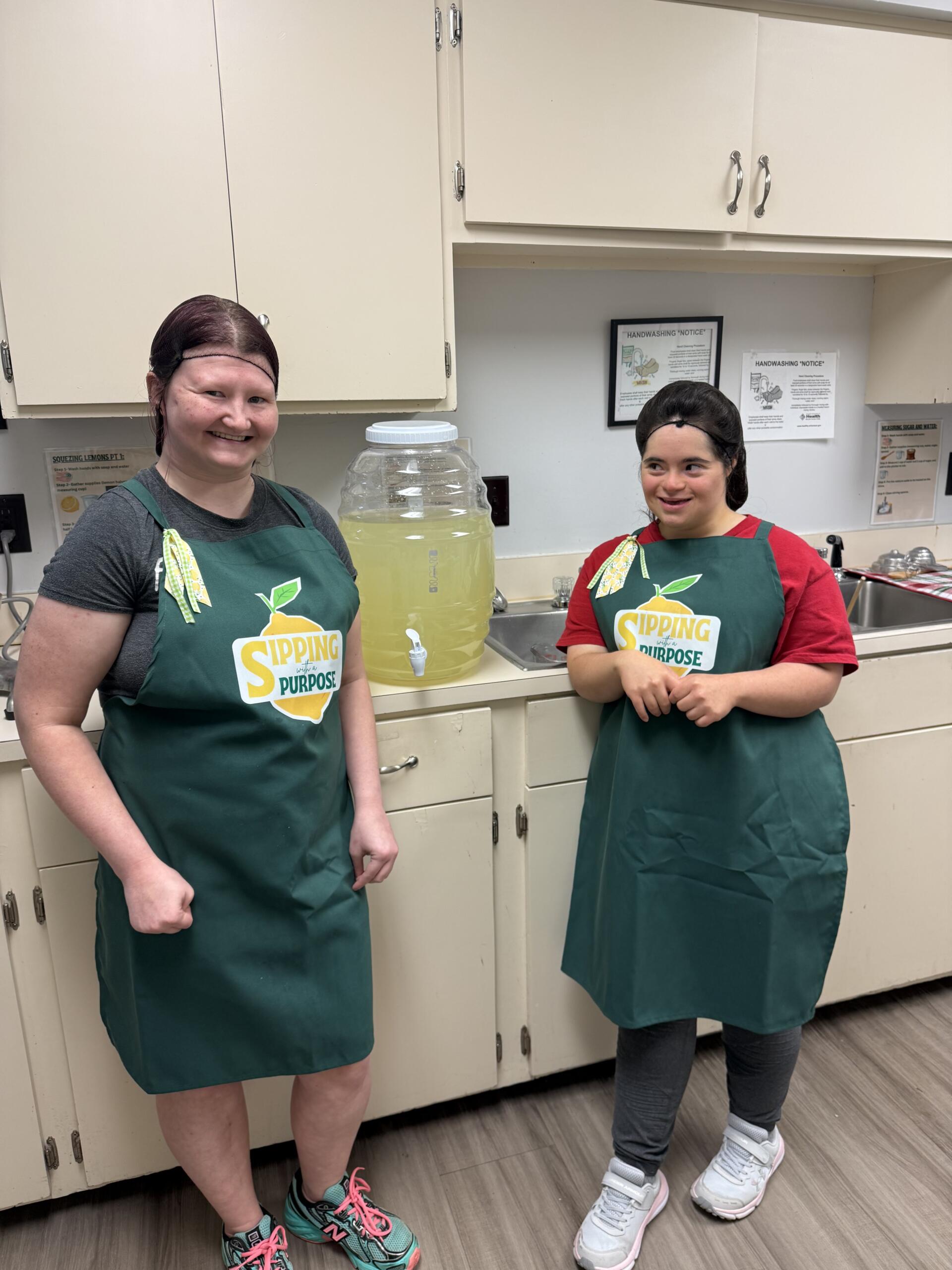Two people in green aprons stand in a kitchen with a large pitcher of lemonade between them, both smiling.