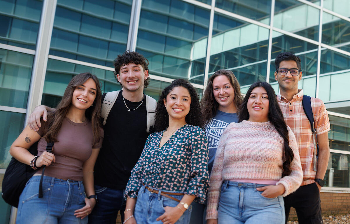 Six diverse students standing together outside a modern glass building, smiling at the camera.