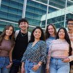 Six diverse students standing together outside a modern glass building, smiling at the camera.