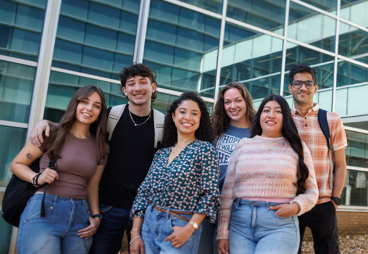 Six diverse students standing together outside a modern glass building, smiling at the camera.