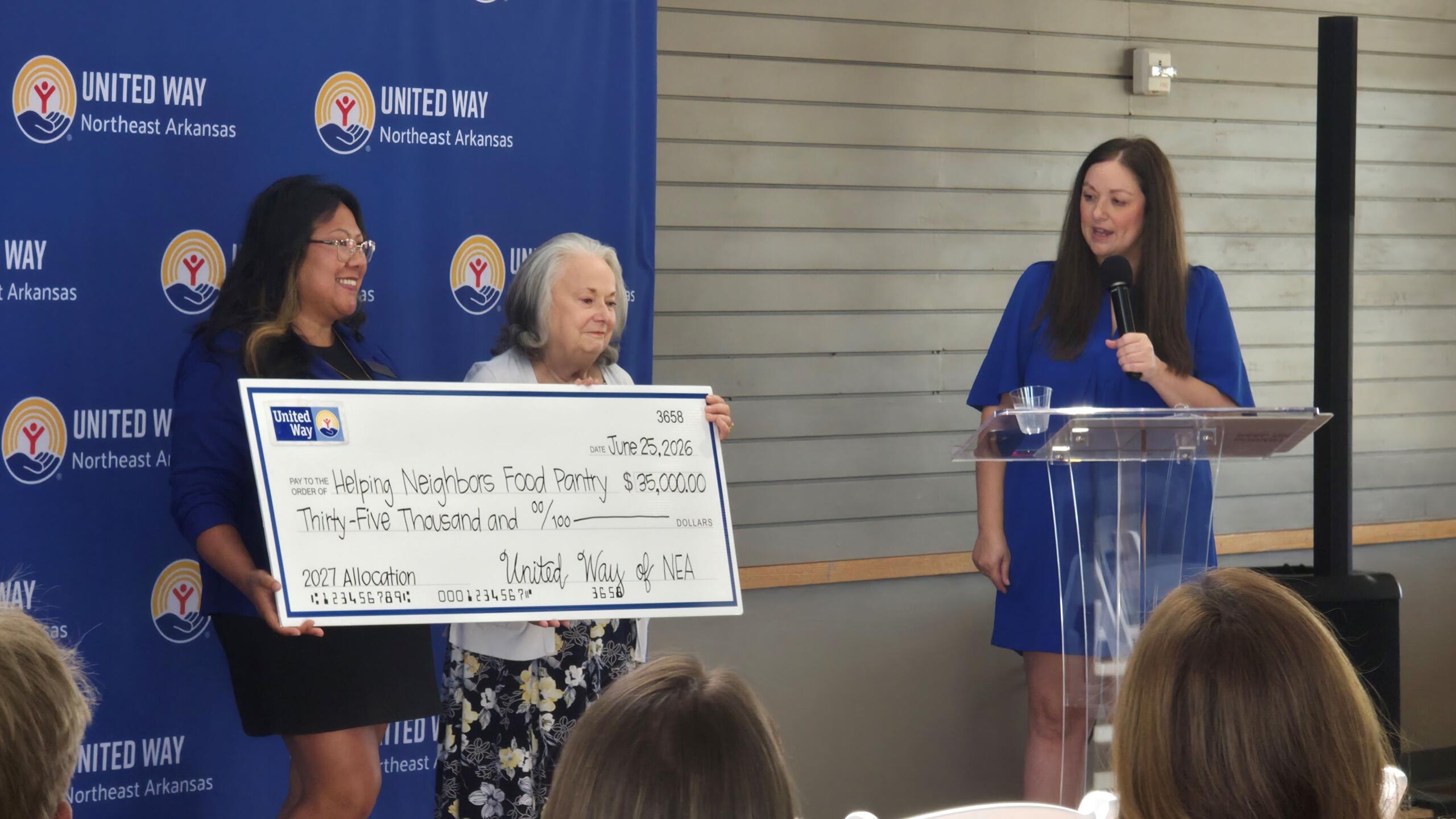 Two women hold a large ceremonial check while a third woman speaks at a podium during a United Way event, with a blue backdrop.