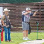 Shooter aims a shotgun at an outdoor range while two men in hats watch nearby. A wooden fence is in the background.