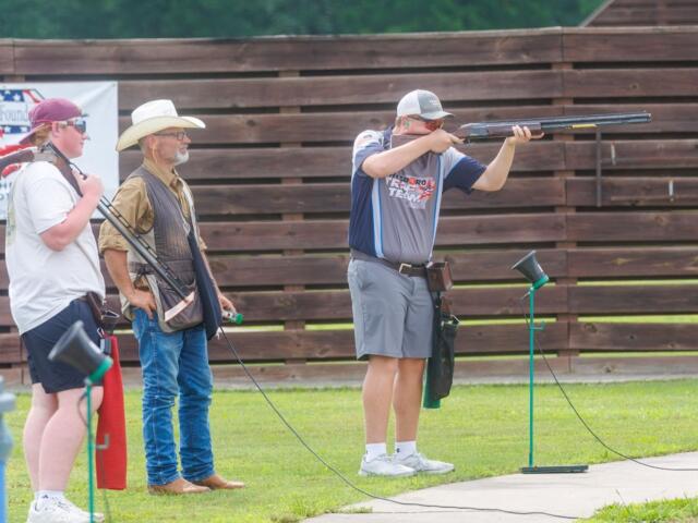 Shooter aims a shotgun at an outdoor range while two men in hats watch nearby. A wooden fence is in the background.